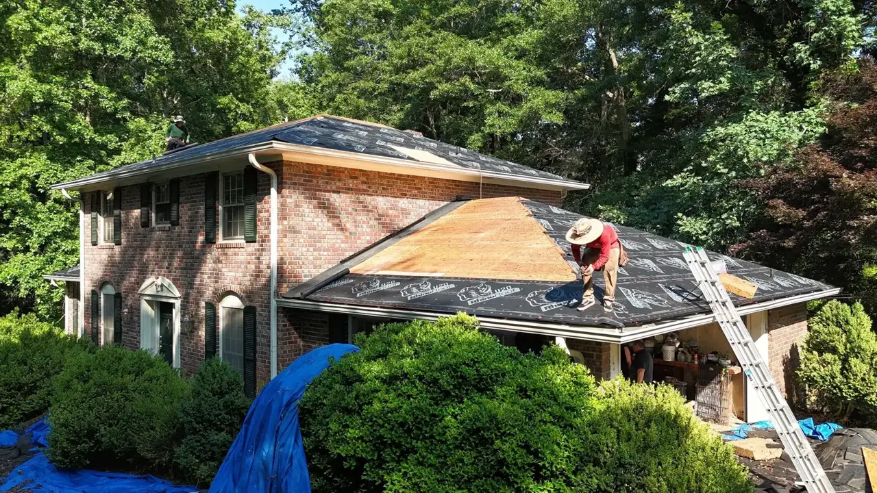 Brick home with partially replaced roof, exposed decking, underlayment, ladders, and worker