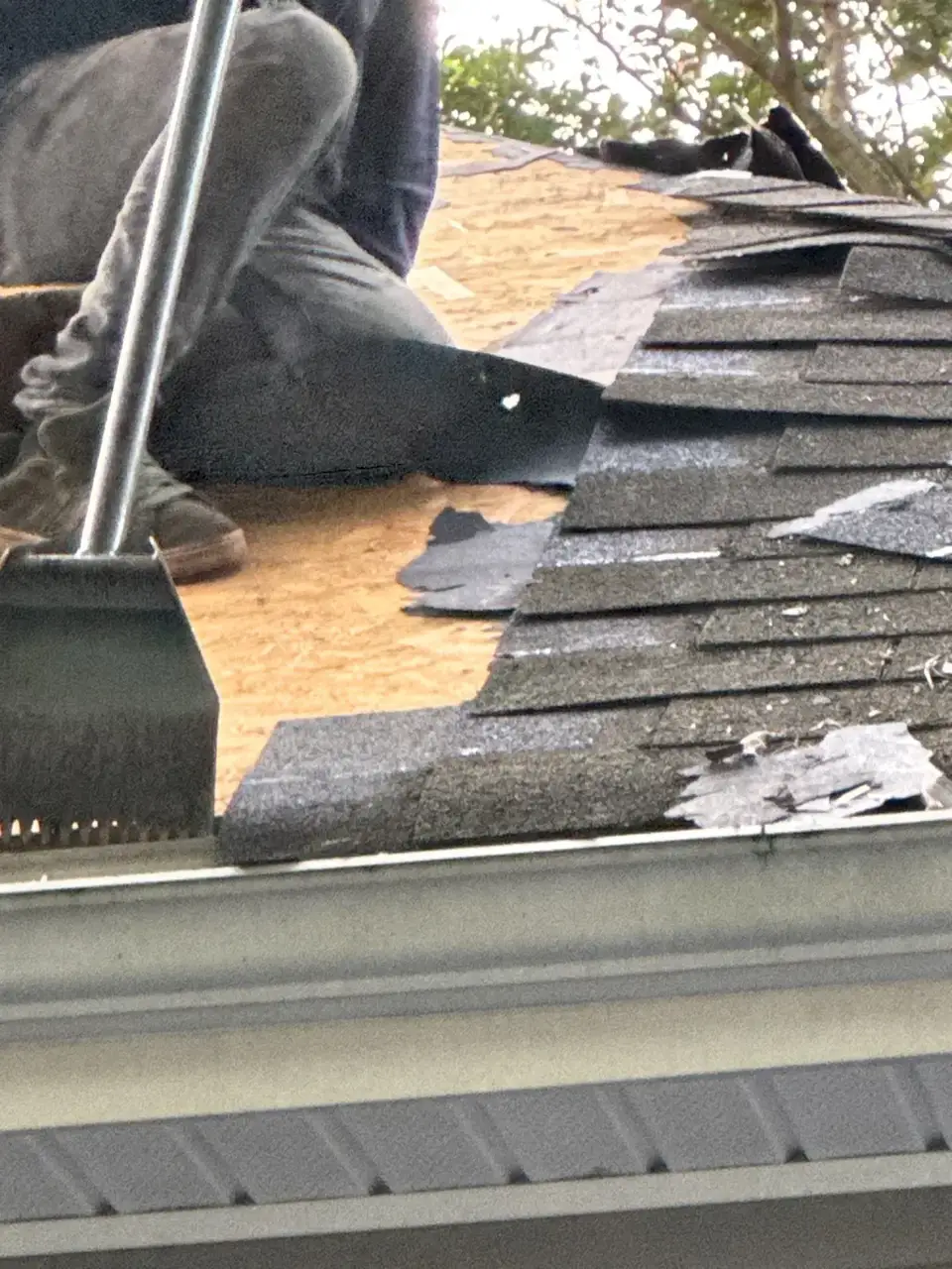 Worker removes roofing shingles beside exposed roof decking and gutter edge.