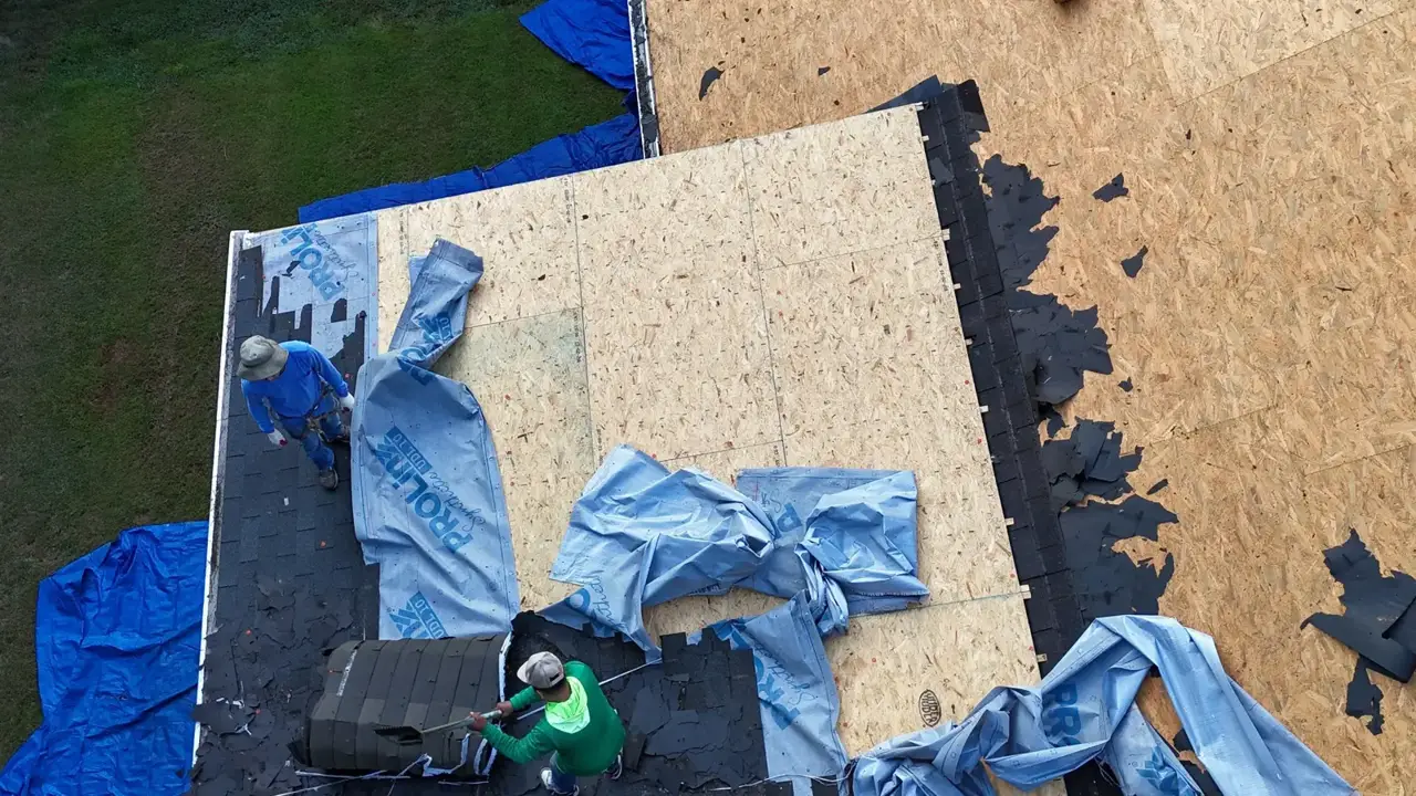 Workers install roofing materials over sheathing, with underlayment rolls and exposed shingles visible in Bethlehem, Georgia.