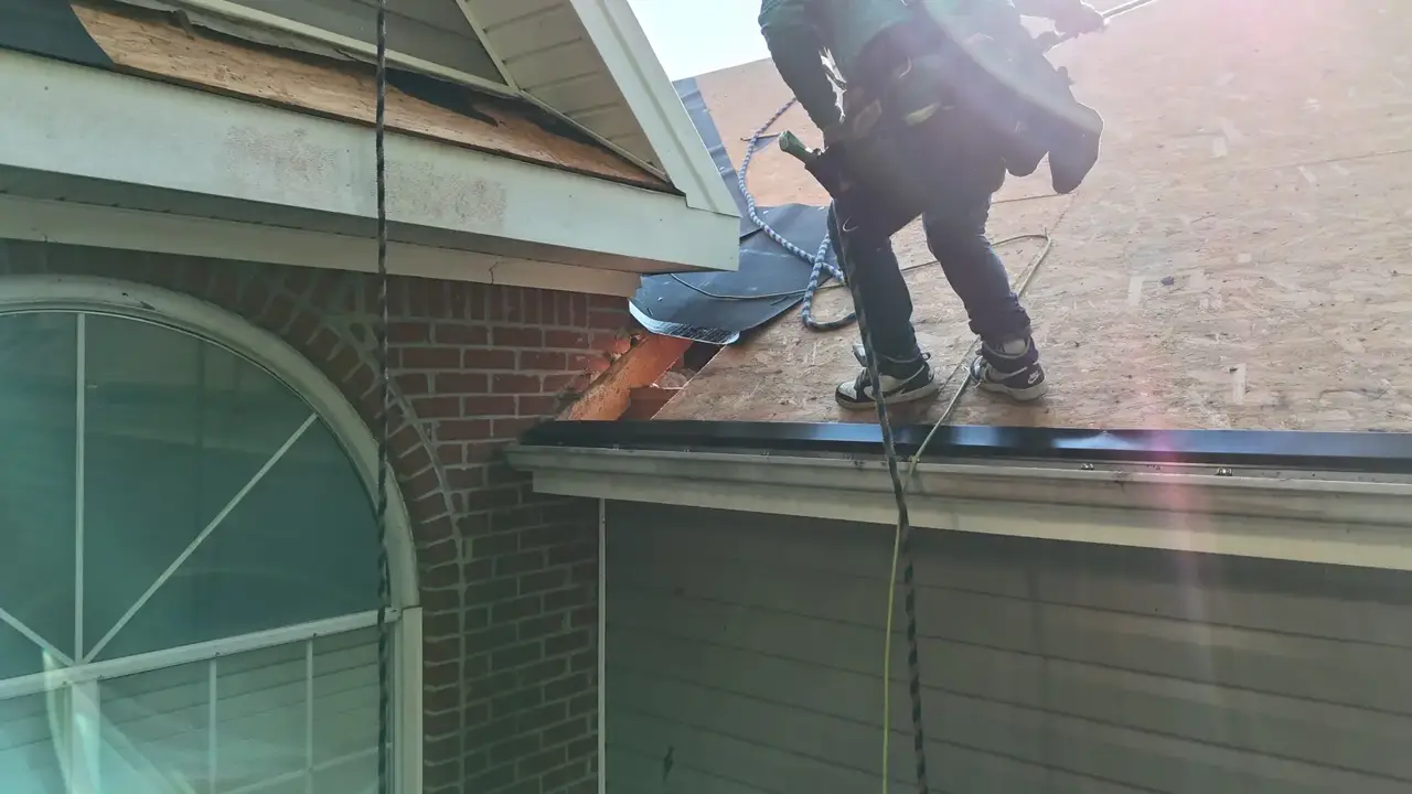 Roofer standing on plywood roof sheathing above a garage with ladder access