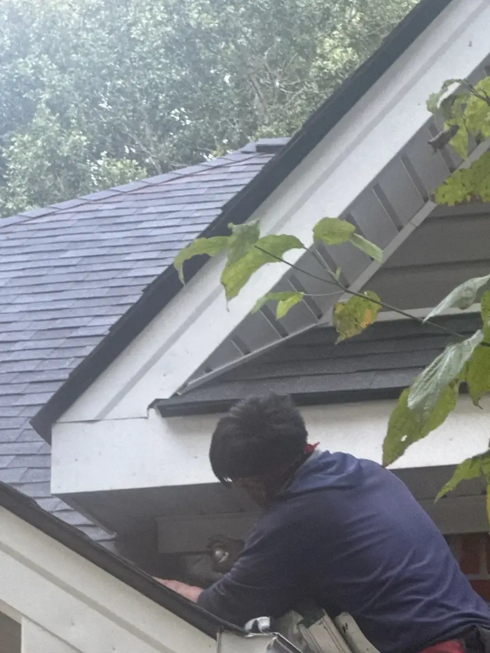 Roofer working beneath a residential roof edge beside gray siding and dark shingles