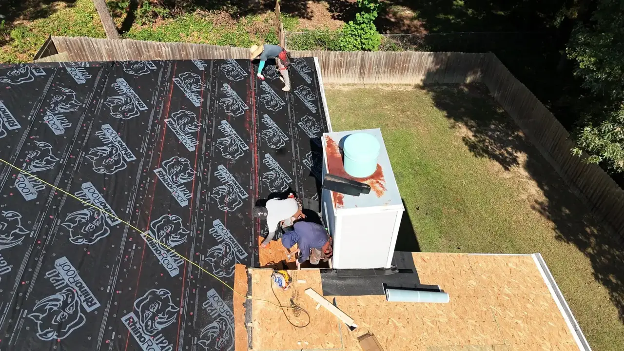 Roof workers stand beside a chimney on plywood decking and black underlayment in Bethlehem, Georgia.