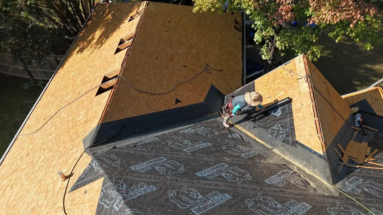 Workers lay black roofing underlayment across newly sheathed roof sections beside exposed plywood decking.