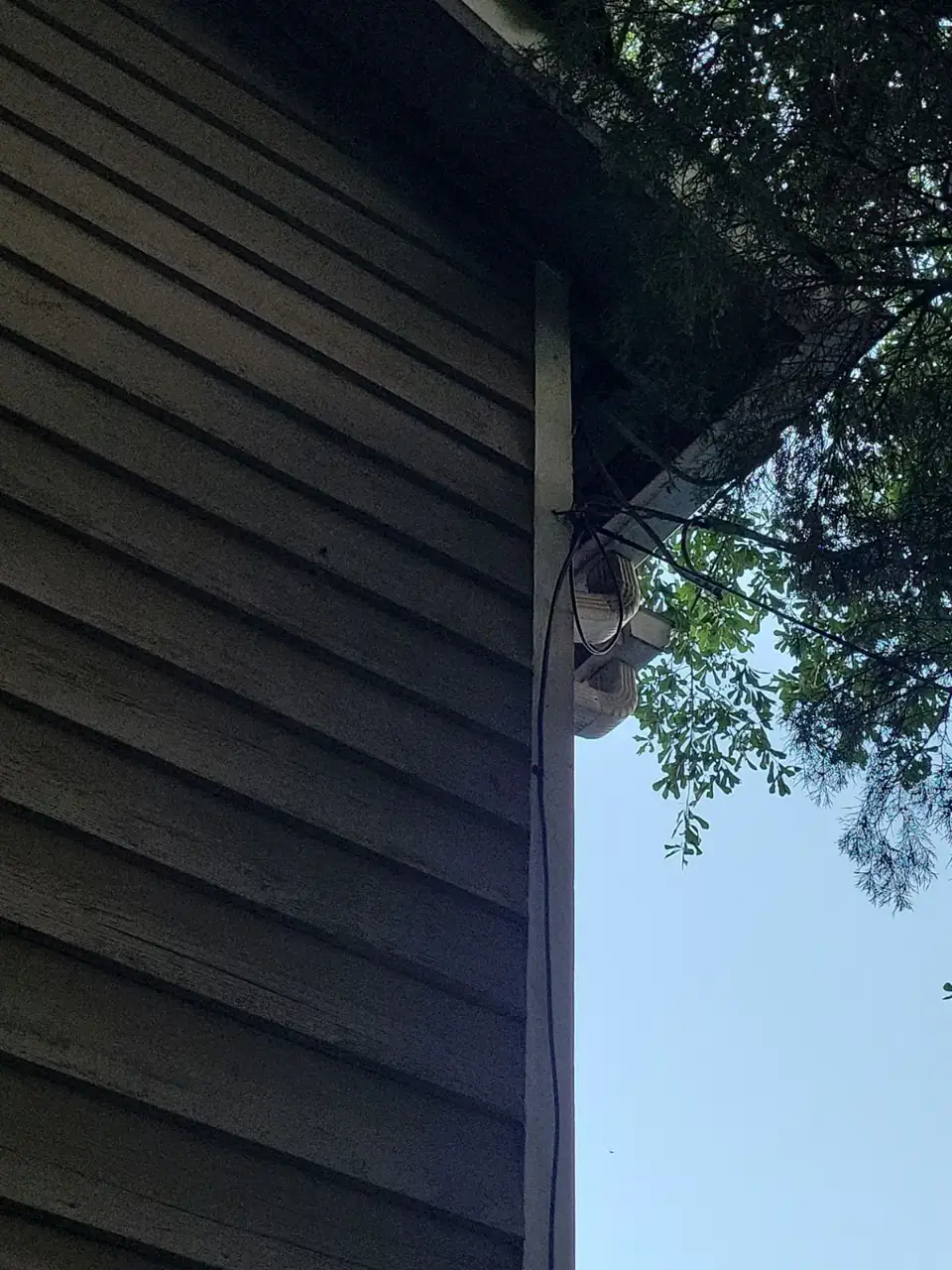 Close view of weathered gray siding, roof eave, wiring, and leafy branches at a Conyers GA home.
