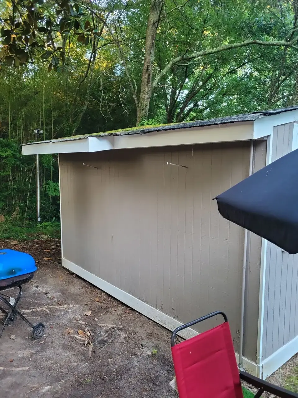 Small outbuilding with weathered low-slope roof, siding, patio furniture, and surrounding trees