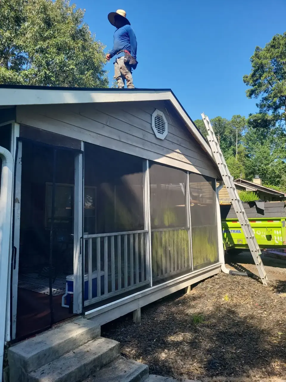 Worker stands on a screened porch roof beside a ladder and green dumpster in Conyers GA.
