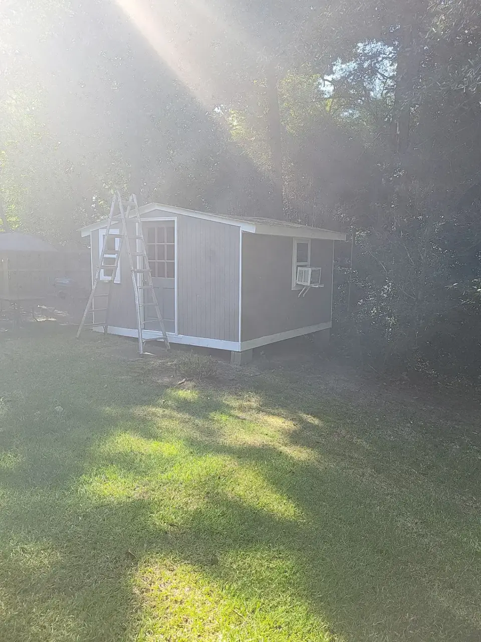 Small gray outbuilding with ladder, window air conditioner, and surrounding grass and trees
