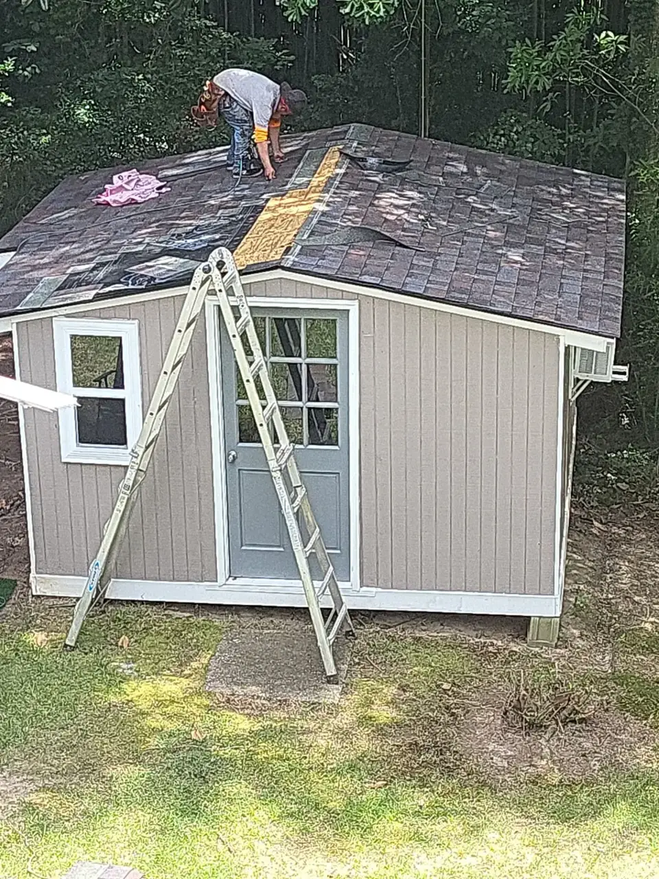 Worker repairs a shed roof beside an extension ladder and exposed roofing underlayment