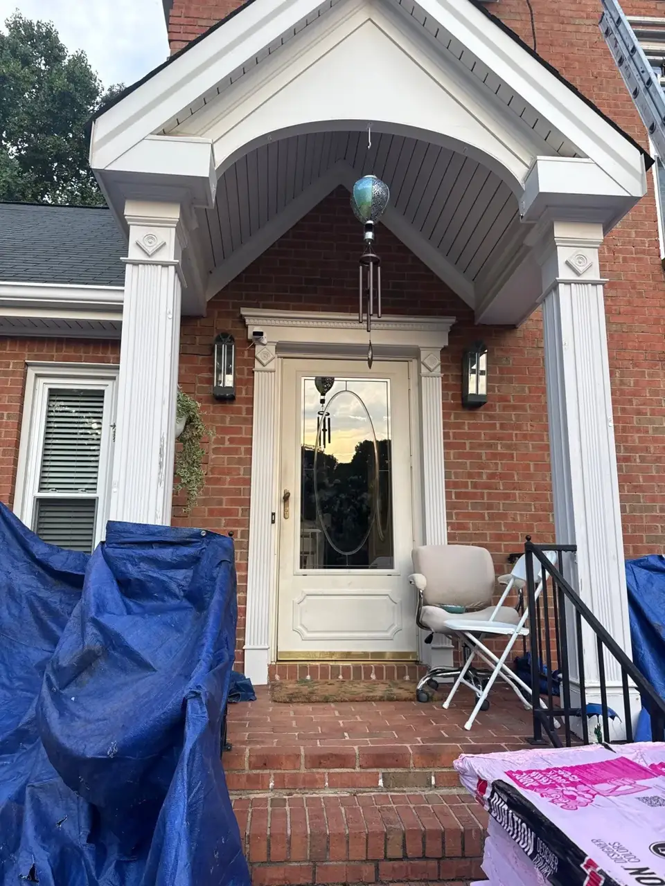 Brick home entrance with covered porch, blue tarps, chairs, and roof edge visible