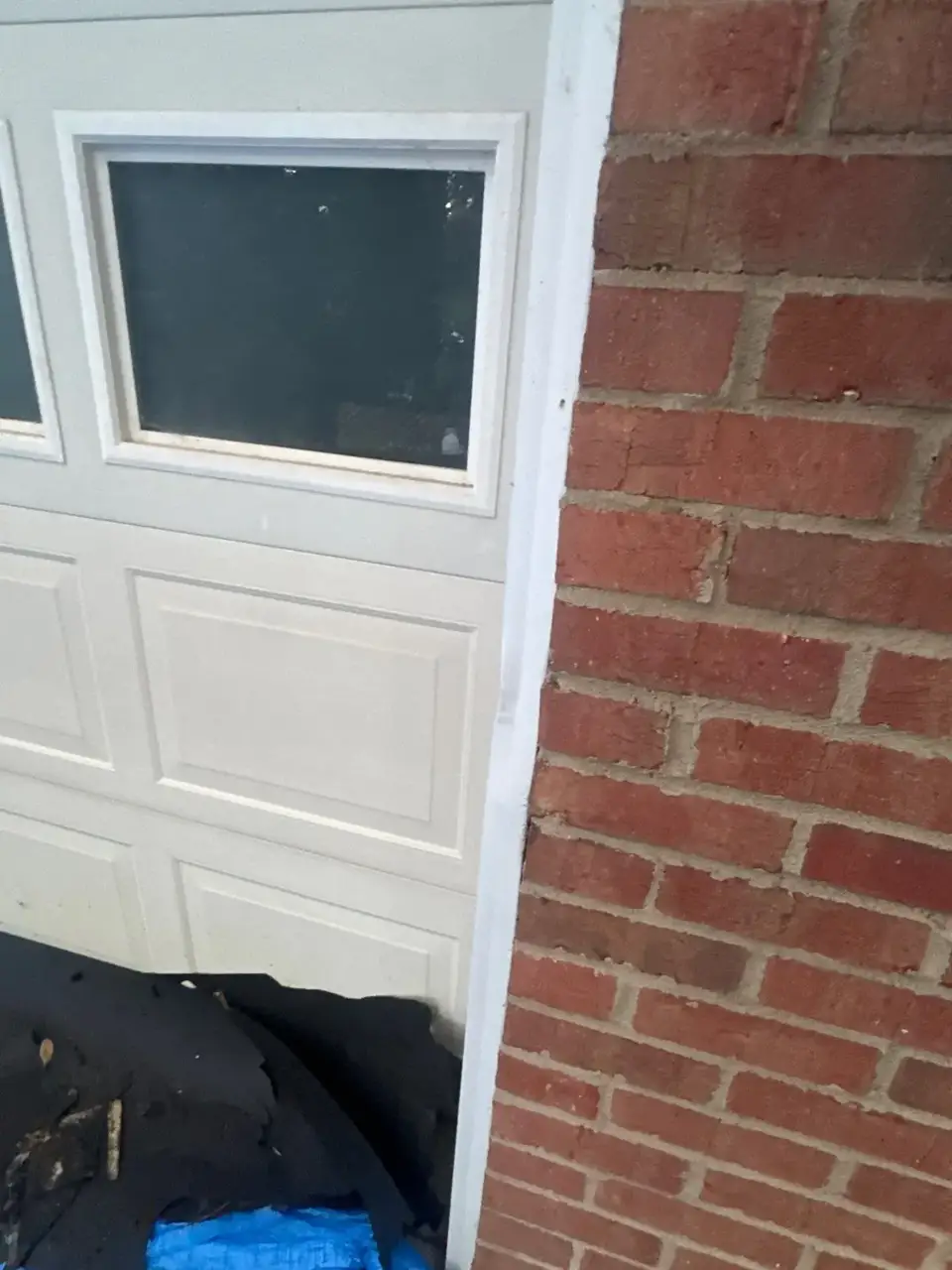Close view of garage door, brick wall, and roofing debris on blue tarp.