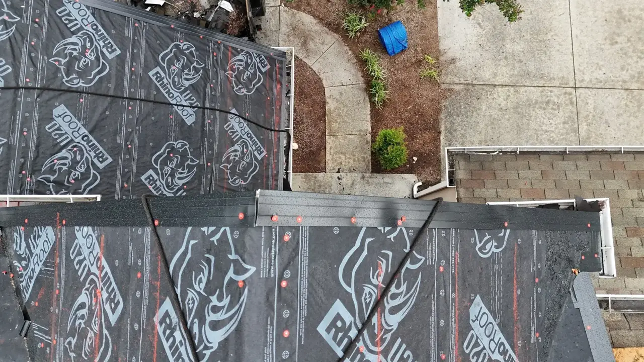 Roof sections covered in black underlayment overlook a concrete walkway, landscaping, and neighboring shingled roofs.
