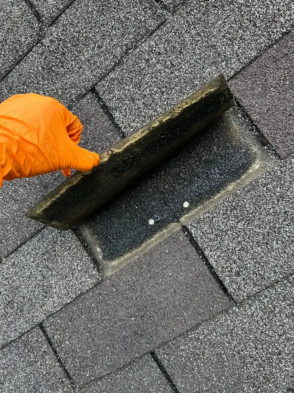 Orange-gloved hand lifts a shingle, revealing exposed underlayment and fasteners beneath.