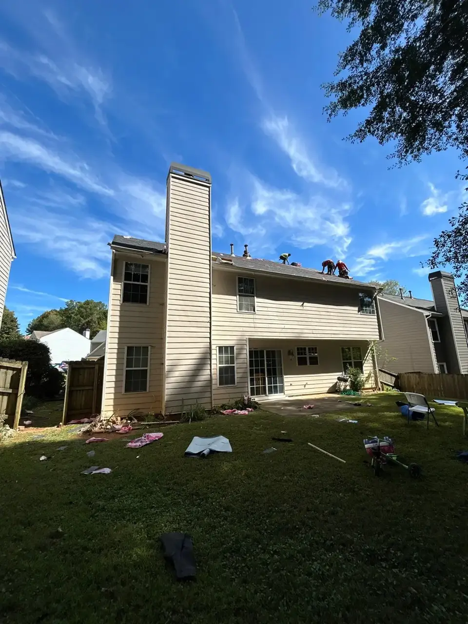 Residential home's rear roof with workers visible on the roof and cleanup debris scattered across the backyard.