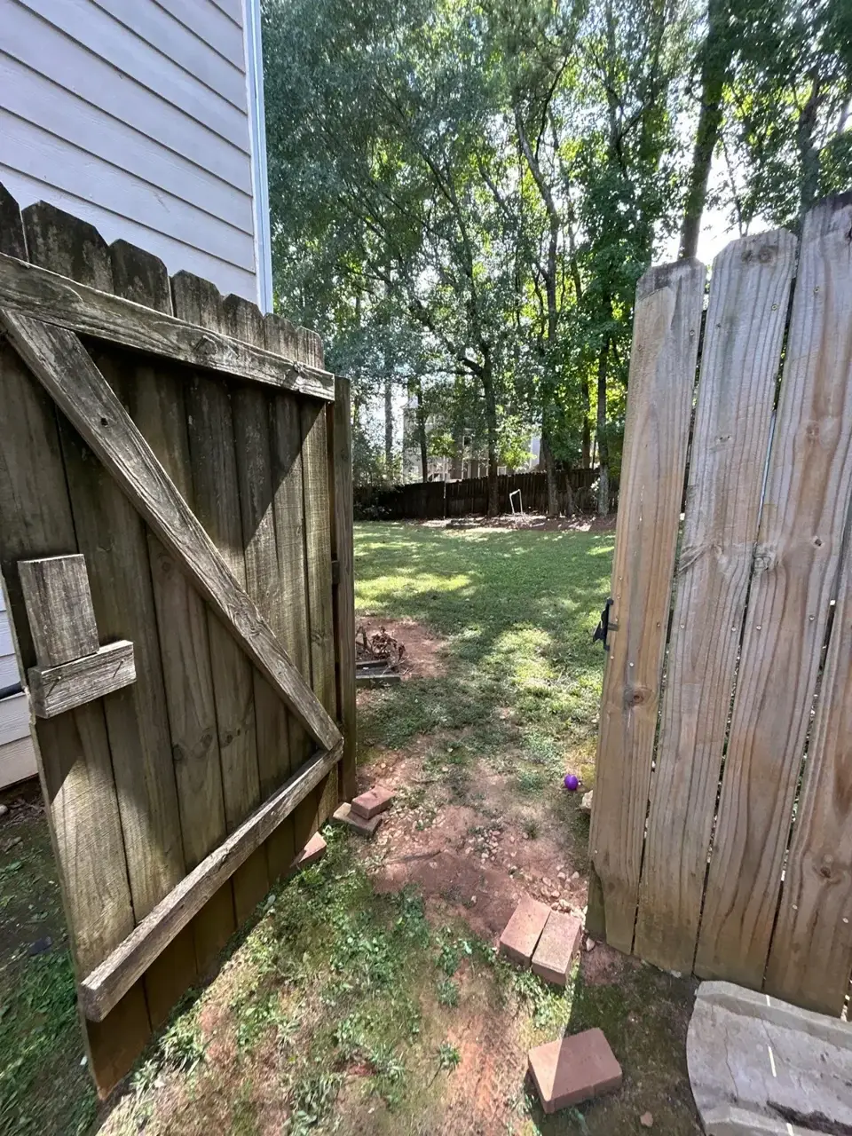 Open wooden backyard gate leading through a fenced residential yard with trees and scattered bricks.