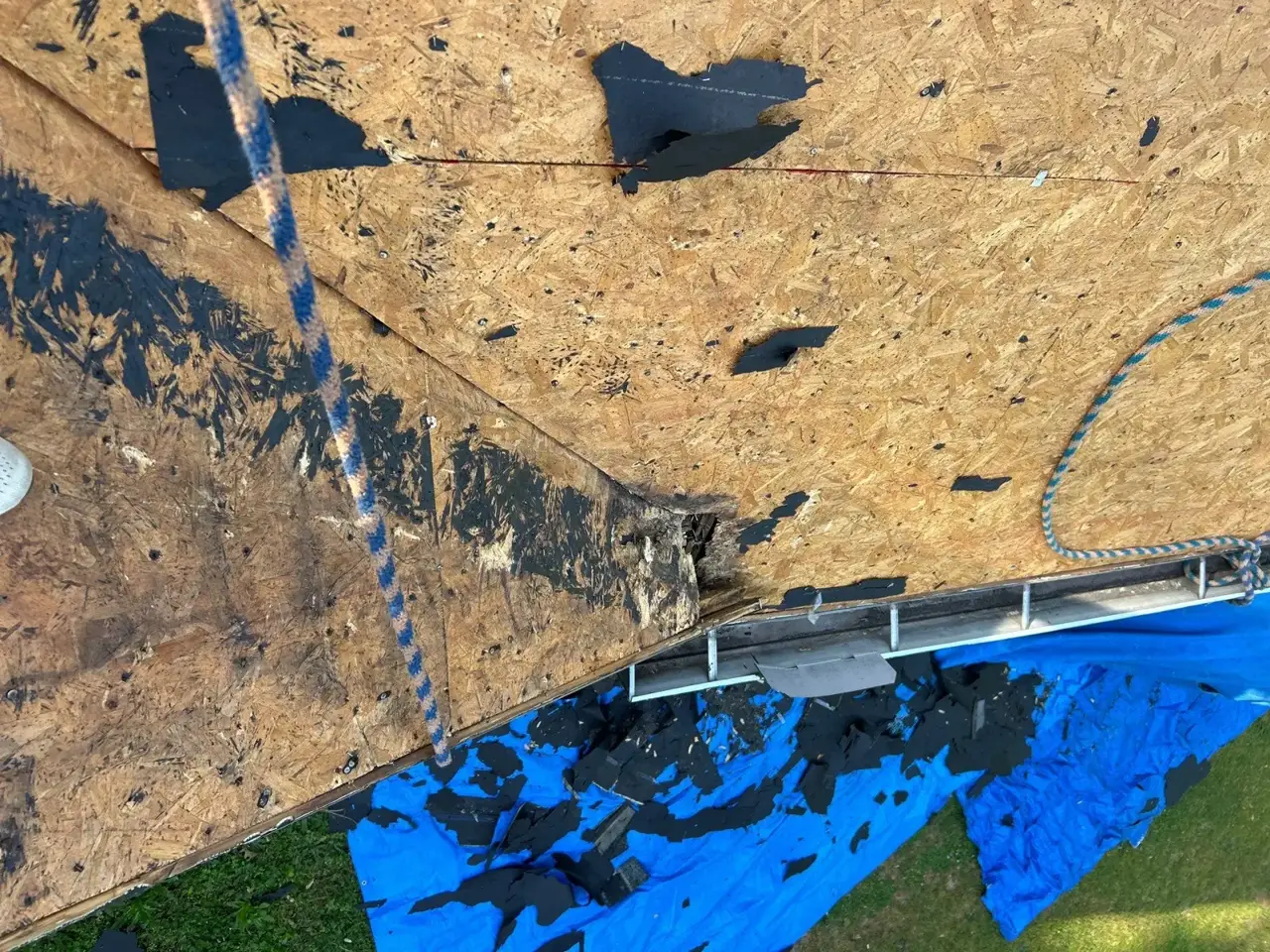 Exposed roof decking with scattered roofing debris, safety rope, gutter, and blue tarp.