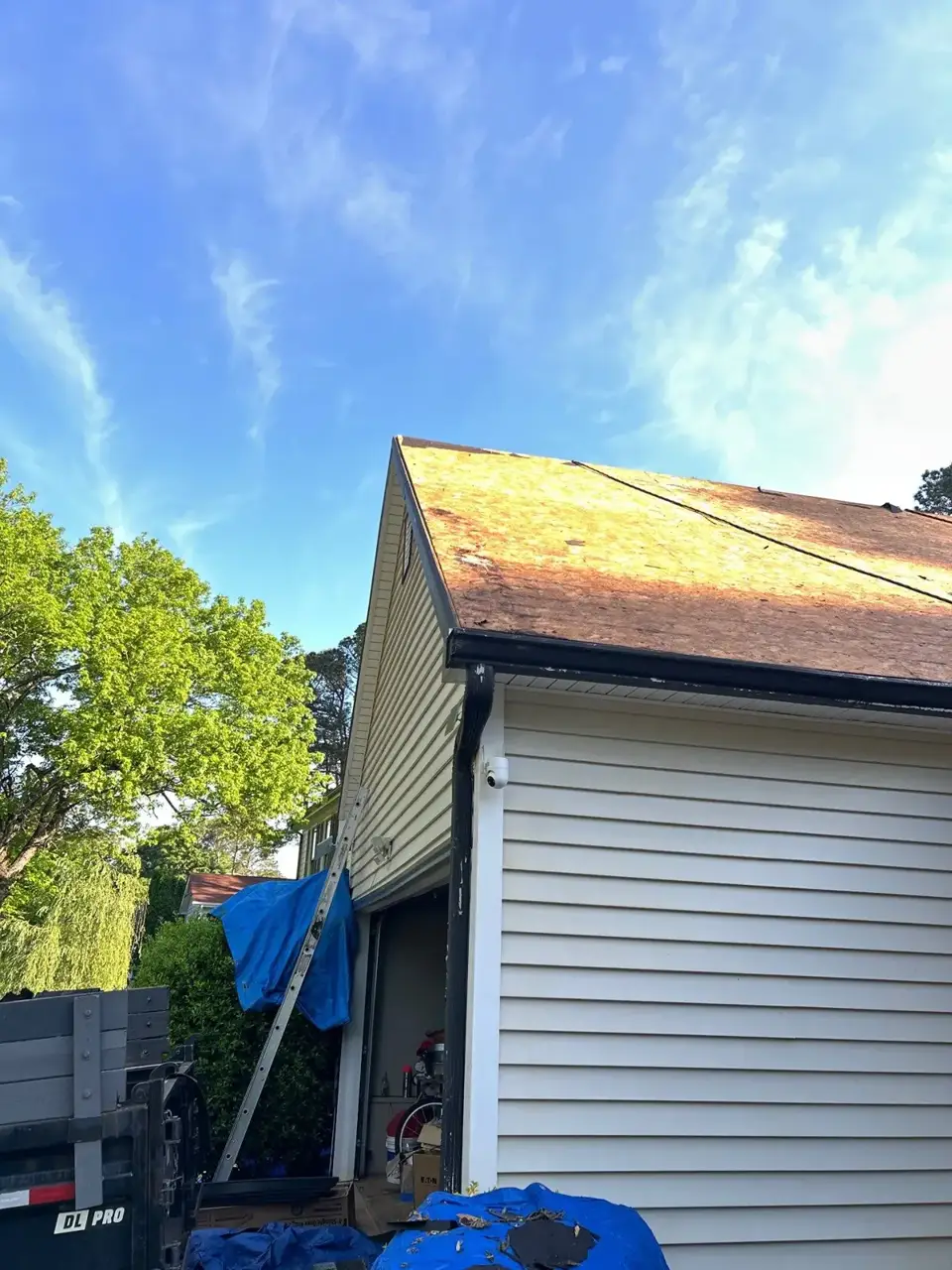 Exposed roof decking above white siding, garage opening, ladder, trailer, and blue tarp.