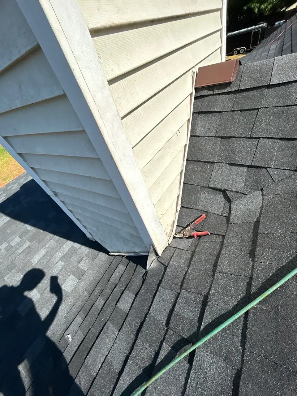 Finished dark shingles beside white siding, with roofing tools and garden hose visible