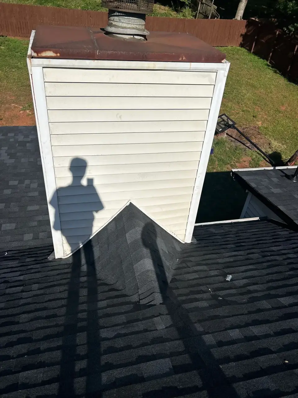 Dark shingle roof surrounds a white-sided chimney with a rusted metal cap and visible shadows.
