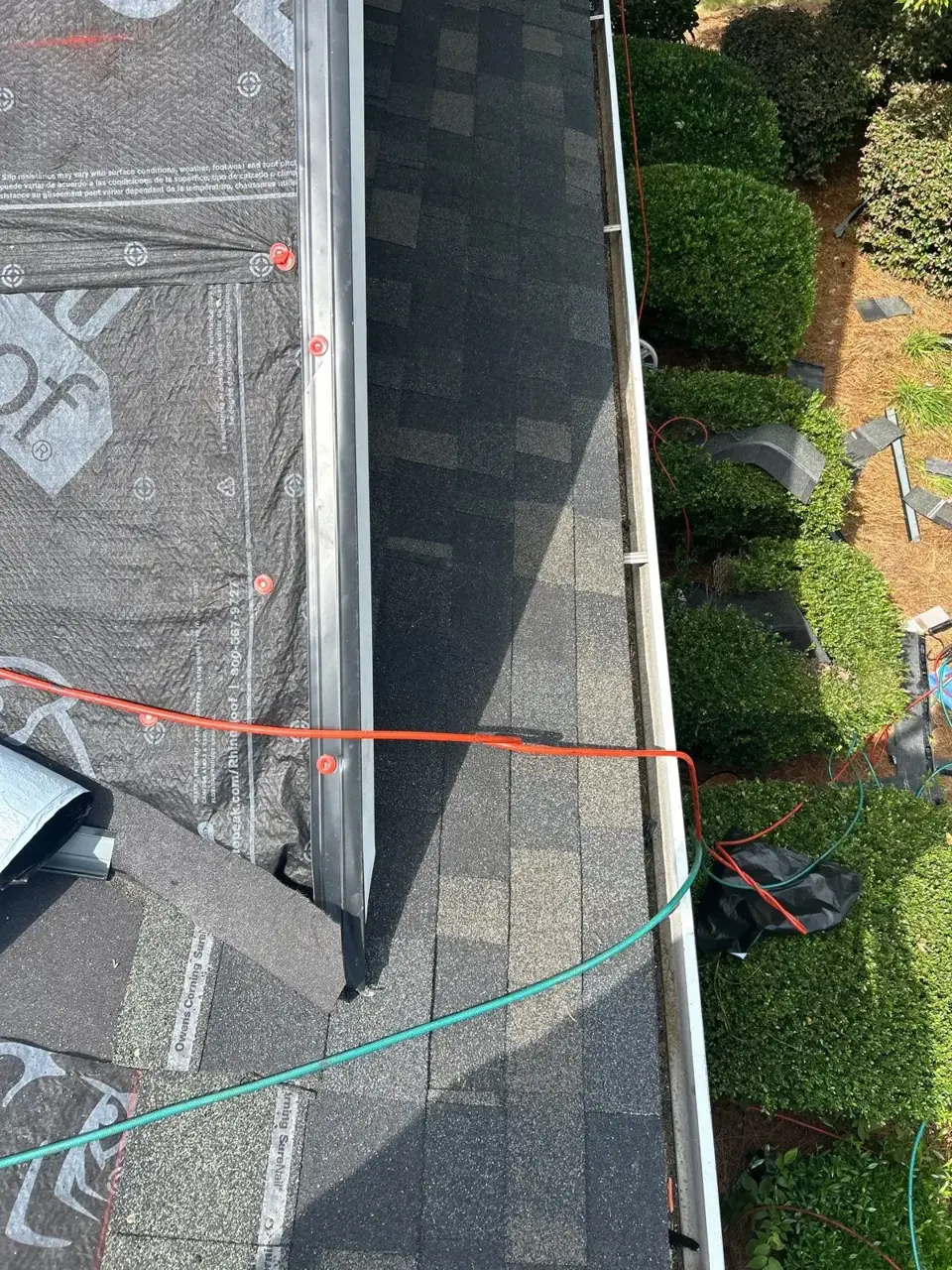 Close view of roof edge shows installed shingles, metal flashing, underlayment, and colored safety hoses.