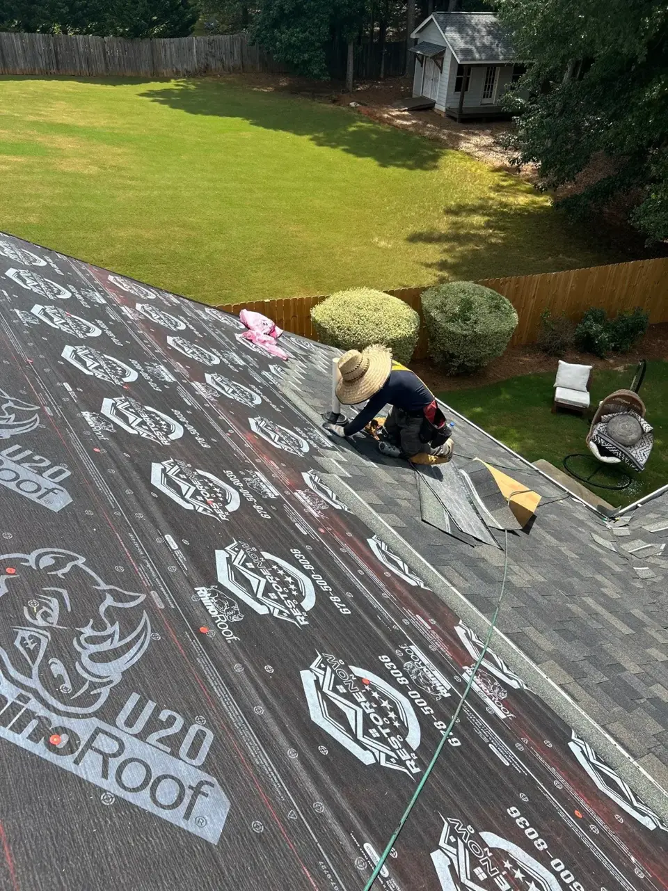 Worker installing roofing materials on underlayment beside completed shingles and backyard landscaping in Loganville, GA