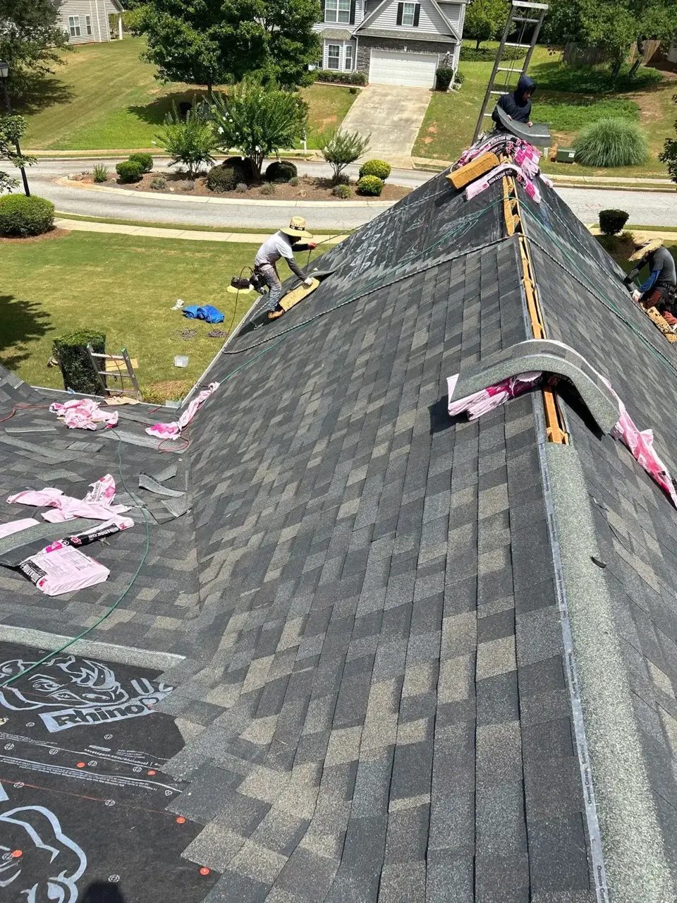 Roofers installing shingles along a residential roof ridge with exposed underlayment and scattered packaging