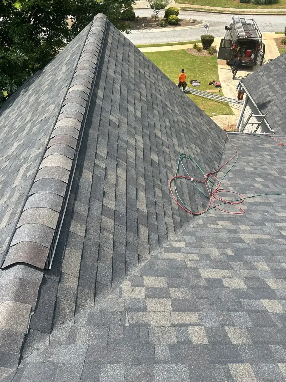 Gray architectural shingle roof with ridge vent, tools, cords, ladder, and worker visible below.