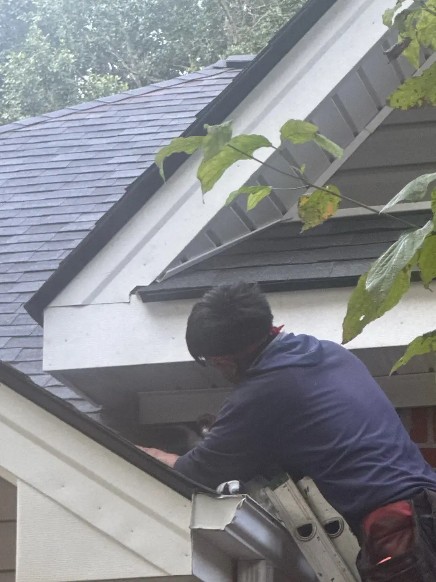 Worker repairing gutter beneath roof eave and leafy branches, Southern Premier Roofing work near Flowery Branch GA